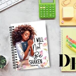 May include: A spiral bound notebook with a white cover featuring a black woman with brown hair, holding a bible. The cover has a floral design and the text "I will not be shaken Psalm 16:8".