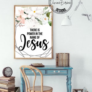May include: A white framed print with a floral design and the text "There is power in the name of Jesus". The print is hanging on a white wall above a blue desk with a wooden chair.