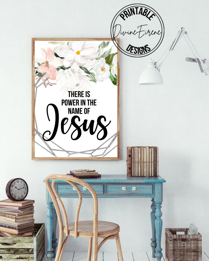 May include: A white framed print with a floral design and the text "There is power in the name of Jesus". The print is hanging on a white wall above a blue desk with a wooden chair.