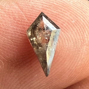 Pode incluir: Uma gema cinza, facetada, em forma de diamante com uma ponta pontiaguda. A gema tem um acabamento áspero e natural e é colocada sobre um fundo rosa claro.