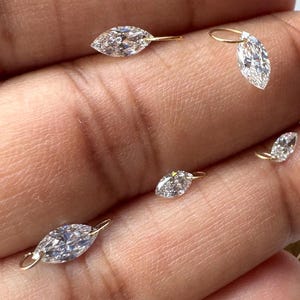 May include: Several marquise-cut diamond charms with gold loops. The diamonds are clear and sparkling, and the gold has a warm, yellow tone. The charms are small and delicate, suitable for jewelry making or embellishments.