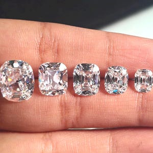 May include: Five clear, cushion-cut gemstones of varying sizes are displayed on a hand. The gems are colorless and highly reflective, showcasing their facets. The largest stone is at the far left, with the sizes decreasing towards the right.