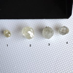 May include: Four rough, uncut diamonds of varying sizes and shapes. The diamonds are a pale, translucent colour, with visible internal structures. They are arranged on a white surface, with a black tray in the background.