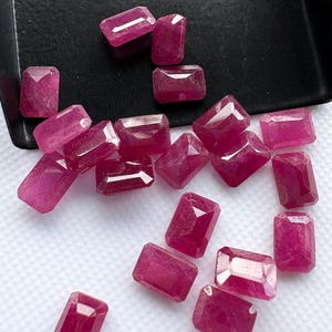 May include: A collection of loose, rectangular-cut rubies with a vibrant pinkish-red hue. These faceted gemstones display a clear, polished surface and are scattered across a white surface, with several stones resting inside a black tray.