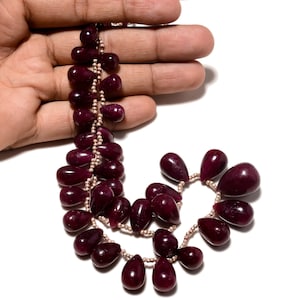 May include: A necklace of polished, teardrop-shaped, deep red gemstones strung on a delicate, light-coloured chain. The gemstones are arranged in a cascading pattern, creating an elegant design. The necklace is displayed against a white background.