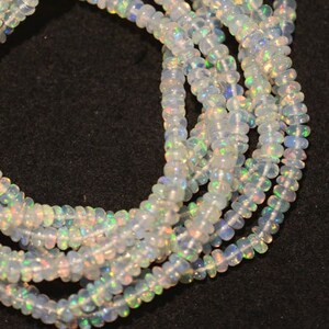 May include: A strand of white opal beads, with iridescent rainbow flashes, strung on a black cord. The beads are small and round, and the strand is long enough to be worn as a necklace or bracelet.