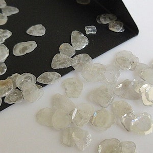 Puede incluir: Un pequeño montón de diamantes en bruto, sin pulir, blancos, también conocidos como diamantes en bruto, que se derraman de una bandeja negra sobre una superficie blanca.