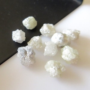 Puede incluir: Primer plano de un peque&ntilde;o mont&oacute;n de diamantes en bruto blancos sobre una superficie blanca. Los diamantes son de diversas formas y tama&ntilde;os.
