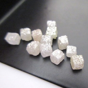 Może przedstawiać: Kolekcja surowych, nieoszlifowanych diamentów. Kamienie szlachetne mają blady, półprzezroczysty kolor z lekko matowym wyglądem. Diamenty mają kształt sześcianu.