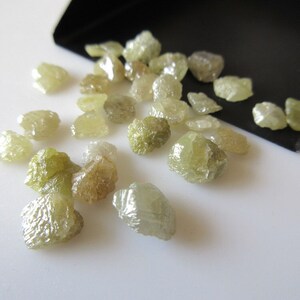 May include: A collection of rough, unpolished, green diamonds. The diamonds are various sizes and shapes, and they are scattered on a white surface.