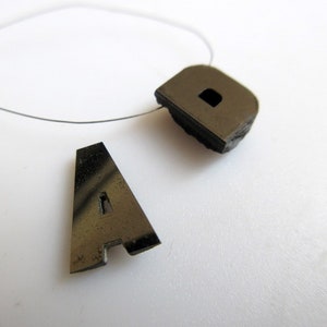 Può includere: Due ciondoli con lettere in metallo grigio scuro, una "A" e una "D", sono mostrati su uno sfondo bianco. Il ciondolo "D" ha un piccolo foro rettangolare. Un filo sottile color argento è attaccato al ciondolo "D".