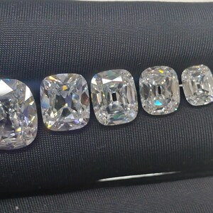 May include: Five cushion-cut diamonds of different sizes are displayed on a black surface. The diamonds are clear and reflect light, showcasing their facets. The largest diamond is on the left, with the others decreasing in size.