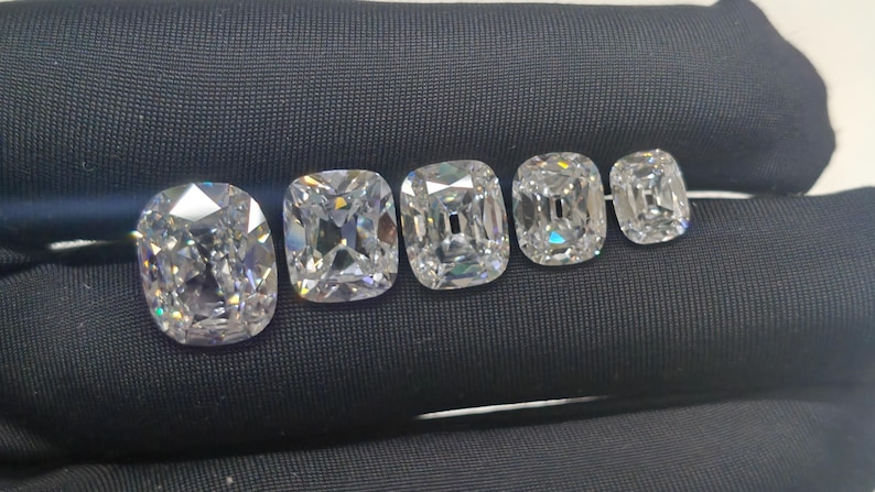 May include: Five cushion-cut diamonds of different sizes are displayed on a black surface. The diamonds are clear and reflect light, showcasing their facets. The largest diamond is on the left, with the others decreasing in size.