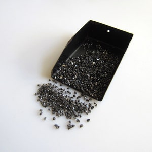 May include: A black metal scoop filled with small, black, shiny, irregularly shaped stones. Some stones have spilled out of the scoop onto a white surface.