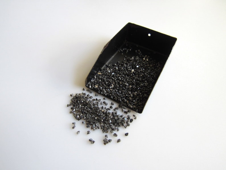 May include: A black metal scoop filled with small, black, shiny, irregularly shaped stones. Some stones have spilled out of the scoop onto a white surface.