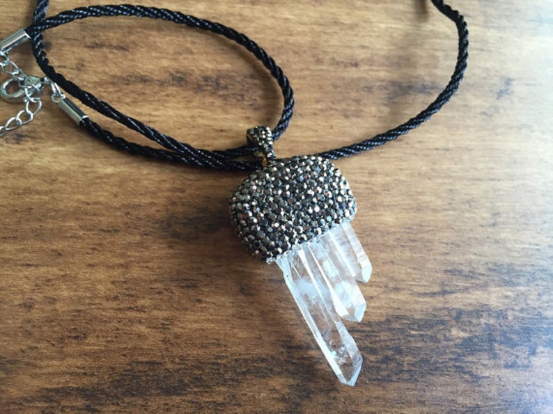 May include: A crystal pendant necklace with a black cord. The pendant features a silver-toned, gem-studded top and three clear quartz crystal points. The necklace is displayed on a wooden surface.