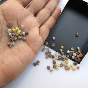 May include: A small pile of rough, uncut diamonds in various colors, including brown, yellow, and gray, are displayed on a hand and spilling out of a black tray.