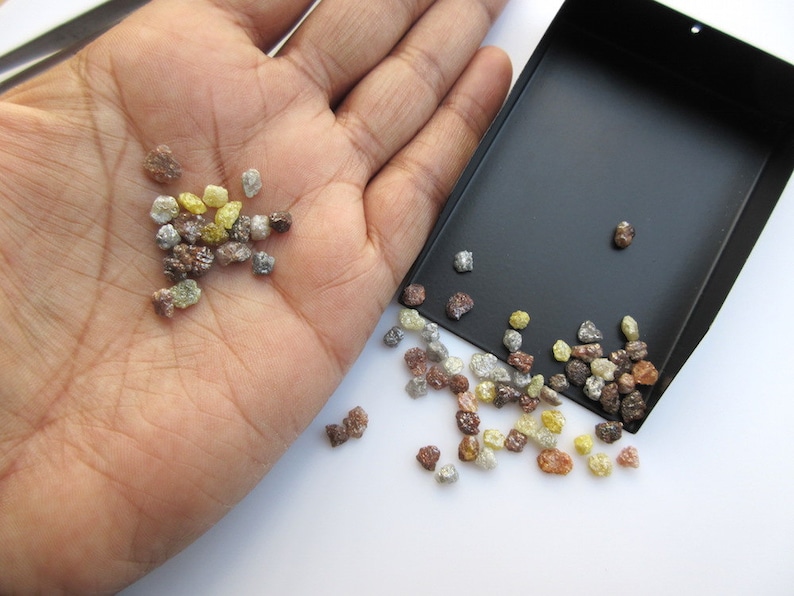 May include: A small pile of rough, uncut diamonds in various colors, including brown, yellow, and gray, are displayed on a hand and spilling out of a black tray.