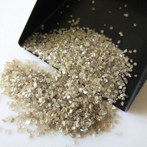 May include: A close-up of a pile of small, rough, light brown diamonds. The diamonds are scattered on a white surface and spilling out of a black scoop.