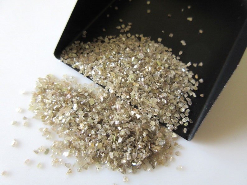 May include: A close-up of a pile of small, rough, light brown diamonds. The diamonds are scattered on a white surface and spilling out of a black scoop.