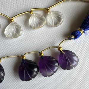 Peut inclure: Un brin de perles de pierres précieuses avec un pompon. Les perles sont sculptées en forme de feuille et alternent entre clair et violet.