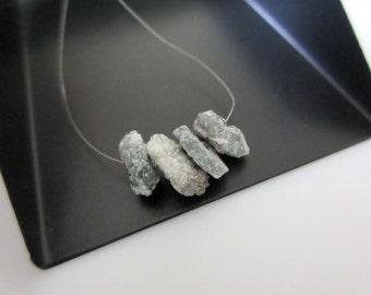 Puede incluir: Cinco piedras preciosas grises verdosas en bruto enhebradas en un hilo transparente. Las piedras preciosas tienen una forma irregular y un ligero brillo metálico.
