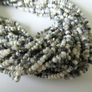 Op de afbeelding: Strengen kleine, ronde kralen in wit, grijs en zwart. De kralen hebben een facetgeslepen oppervlak, waardoor een textuur ontstaat. De kralen zijn aan elkaar geregen en vormen een ketting of armband.