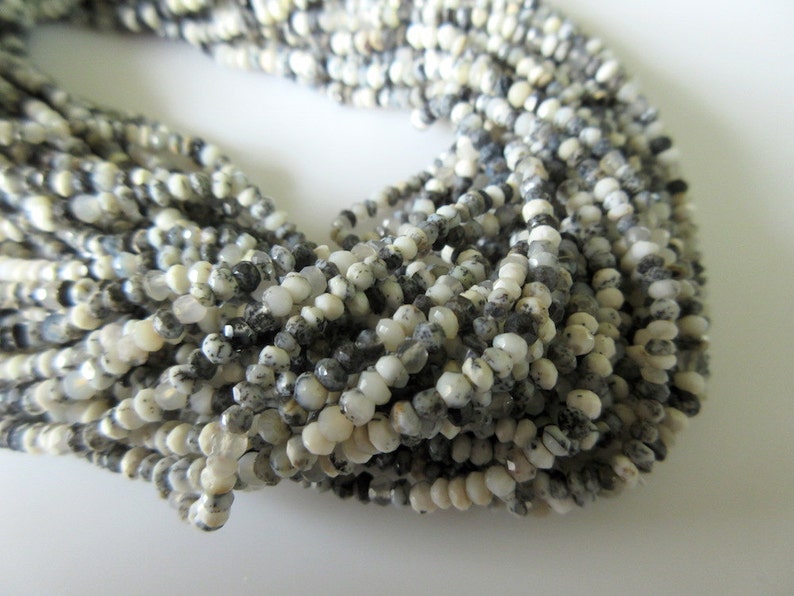 Op de afbeelding: Strengen kleine, ronde kralen in wit, grijs en zwart. De kralen hebben een facetgeslepen oppervlak, waardoor een textuur ontstaat. De kralen zijn aan elkaar geregen en vormen een ketting of armband.