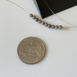 May include: A necklace featuring small, round, gray beads strung on a thin wire. A U.S. quarter coin is next to the necklace for size comparison. The beads have a speckled appearance. The background is white.