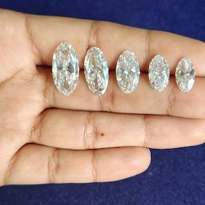 May include: Five loose, clear, colorless, oval-cut gemstones arranged in a row by size from largest to smallest. Each stone features a faceted surface that reflects light, creating a brilliant, sparkling appearance against the dark blue background.