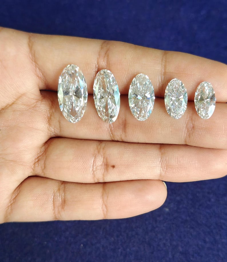May include: Five loose, clear, colorless, oval-cut gemstones arranged in a row by size from largest to smallest. Each stone features a faceted surface that reflects light, creating a brilliant, sparkling appearance against the dark blue background.