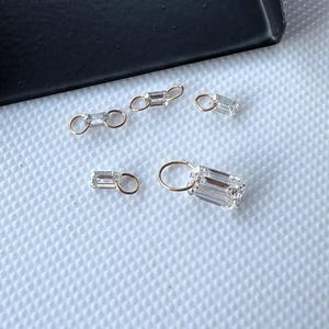 May include: Close-up of several small, gold-colored jewelry pieces with clear, rectangular-cut gemstones. The pieces are scattered on a white, textured surface, with a black edge in the background. The gemstones have a brilliant, faceted appearance.
