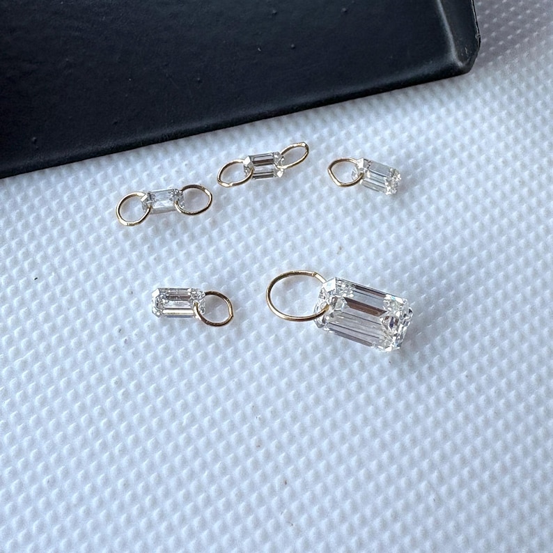 May include: Close-up of several small, gold-colored jewelry pieces with clear, rectangular-cut gemstones. The pieces are scattered on a white, textured surface, with a black edge in the background. The gemstones have a brilliant, faceted appearance.