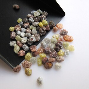 May include: A small scoop filled with a variety of rough, uncut diamonds. The diamonds are in shades of brown, gray, yellow, and clear. The diamonds are spilling out of the scoop onto a white surface.