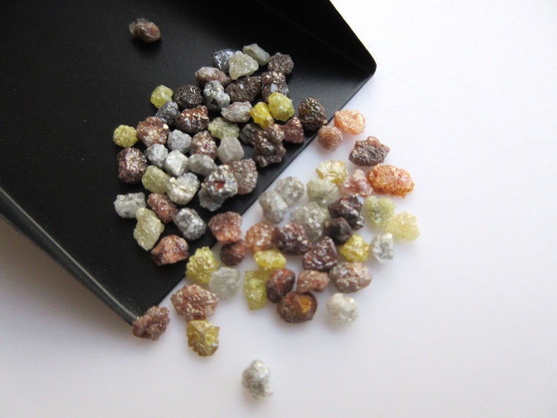 May include: A small scoop filled with a variety of rough, uncut diamonds. The diamonds are in shades of brown, gray, yellow, and clear. The diamonds are spilling out of the scoop onto a white surface.