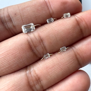 May include: A collection of small, clear, rectangular-cut gemstone earrings with gold-colored hoops. The earrings are displayed against a neutral background, showcasing their sparkle and design. The gemstones are set in a minimalist style.