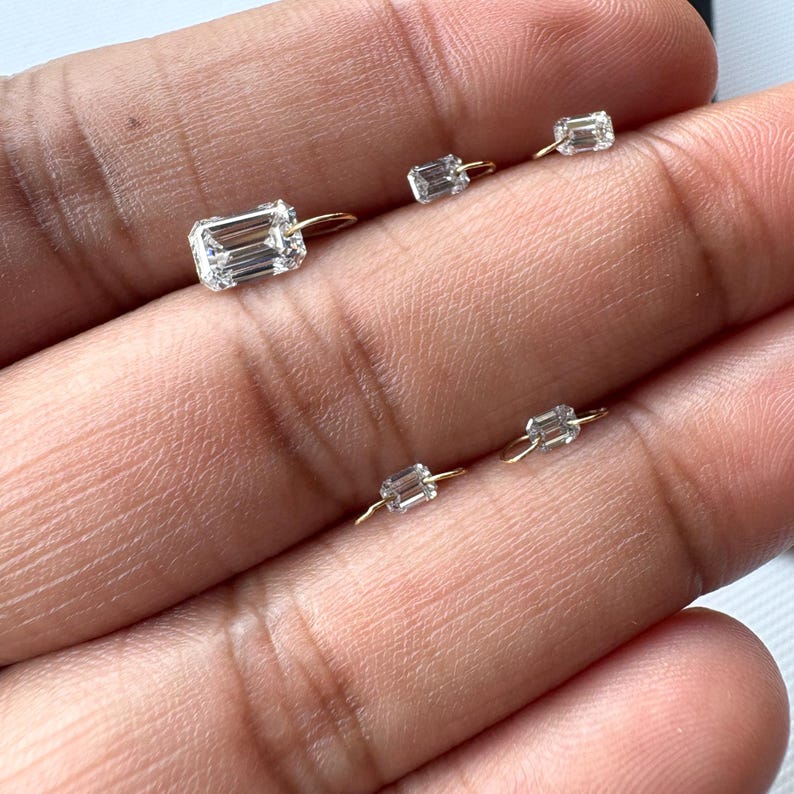May include: A collection of small, clear, rectangular-cut gemstone earrings with gold-colored hoops. The earrings are displayed against a neutral background, showcasing their sparkle and design. The gemstones are set in a minimalist style.