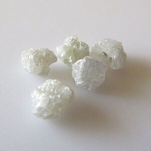 Puede incluir: Cinco cristales de diamante en bruto, blancos, sobre una superficie blanca.