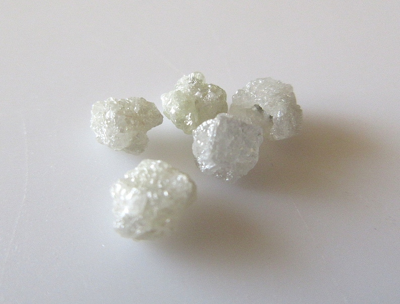 Puede incluir: Cinco cristales de diamante en bruto, blancos, sobre una superficie blanca.