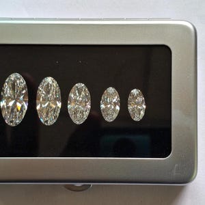 May include: A rectangular silver-colored metal display case with a hinged lid and a clear window. Inside, five clear, faceted, oval-shaped gemstones are arranged in a row on a black velvet-like insert, decreasing in size from left to right.