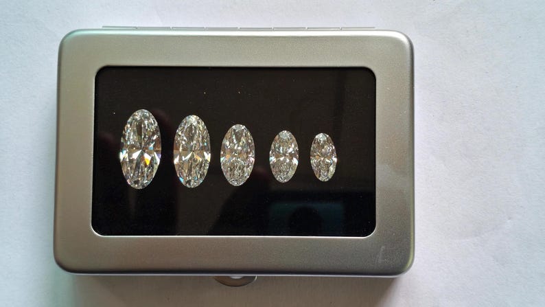 May include: A rectangular silver-colored metal display case with a hinged lid and a clear window. Inside, five clear, faceted, oval-shaped gemstones are arranged in a row on a black velvet-like insert, decreasing in size from left to right.