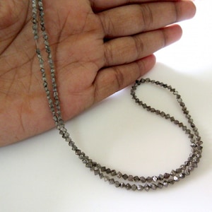 May include: A strand of grey diamond beads, strung on a thin wire, is displayed on a hand. The beads are small and have a faceted, rough-cut appearance.
