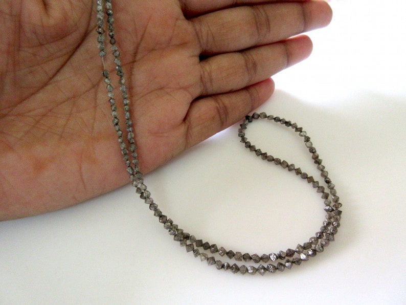May include: A strand of grey diamond beads, strung on a thin wire, is displayed on a hand. The beads are small and have a faceted, rough-cut appearance.
