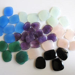 May include: Assortment of smooth, polished gemstone cabochons in various colours, including blue, green, purple, pink, and black. The stones are arranged in a semi-circle on a white background.