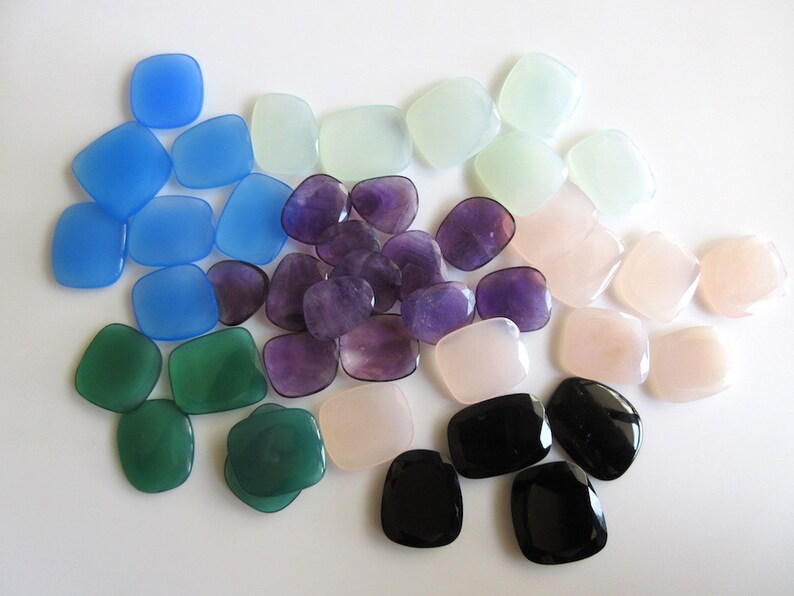 May include: Assortment of smooth, polished gemstone cabochons in various colours, including blue, green, purple, pink, and black. The stones are arranged in a semi-circle on a white background.