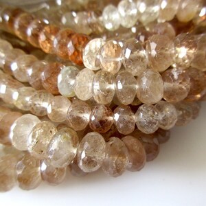May include: A strand of faceted, translucent brown gemstone beads. The beads are round and have a slightly irregular shape. The beads are strung on a clear thread.
