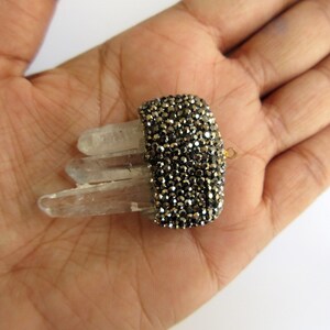 May include: A crystal quartz ring with a rectangular, dark gray, gem-studded band. The ring features three clear quartz crystals. The ring is held in a hand against a white background.