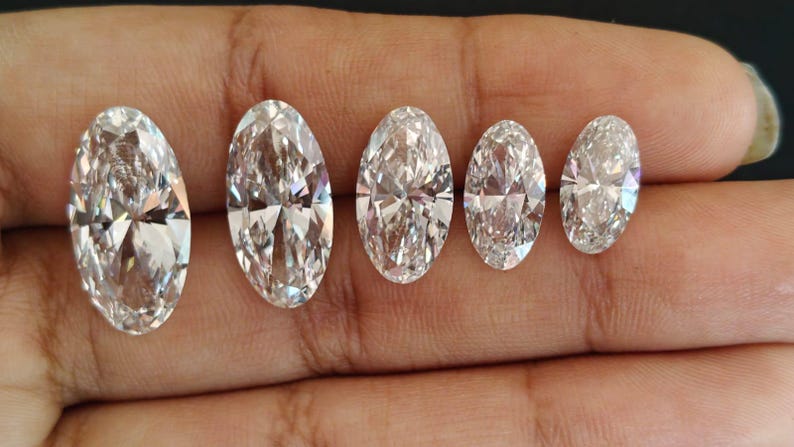 May include: Five clear, colorless, oval-cut gemstones arranged in a row by size from largest to smallest. Each stone features a faceted surface that reflects light, creating a brilliant, sparkling appearance with sharp, geometric edges.