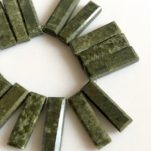 May include: A collection of 14 green jade stone rectangular beads with faceted edges. The beads are arranged in a circle on a white surface.