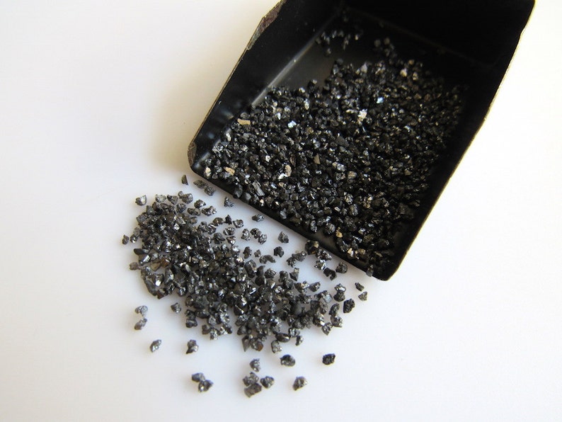 May include: A small black metal scoop filled with black diamond grit. The grit is spilling out of the scoop onto a white surface.